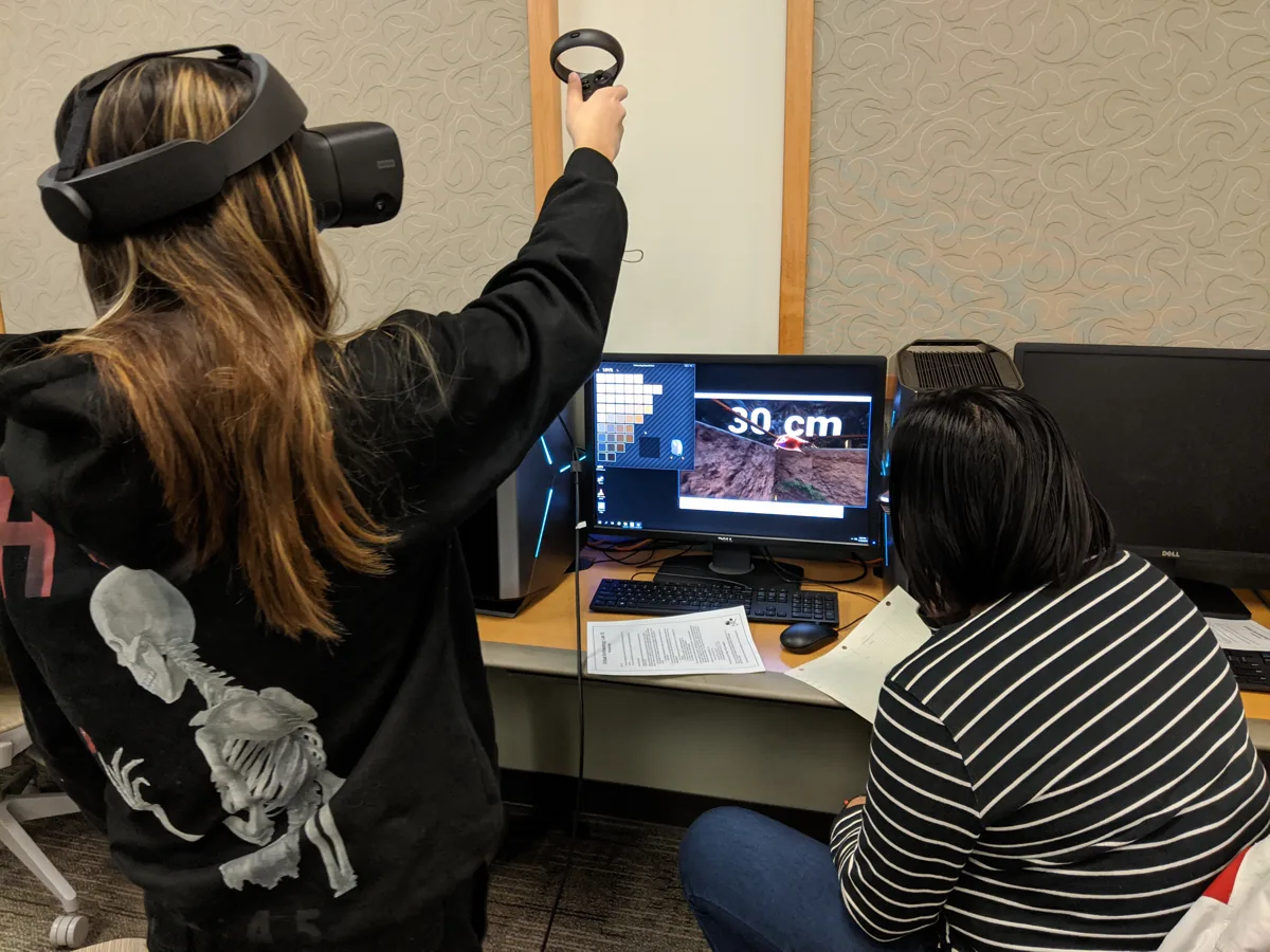 A participant holds a VR controller up while another person sits beside the workstation.